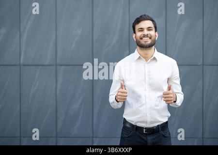 Ein lächelnder Mann mit dunklen Haaren und Bart zeigt die Daumen hoch, gekleidet in einem weißen Hemd. Der Hintergrund ist eine graue strukturierte Wand. Stockfoto