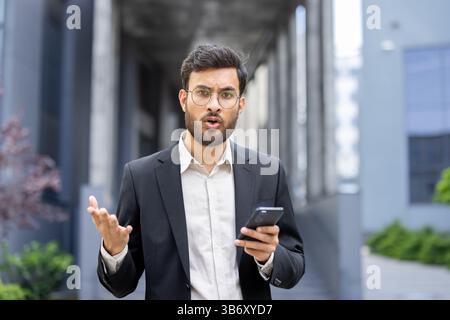 Ein Geschäftsmann, der in einem Anzug gekleidet ist, sieht überrascht und animiert aus, während er ein Telefon hält und mit seinen Händen im Freien Gesten bewegt. Stockfoto