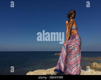 Schöne Frau in langem, farbenfrohen Rock, die am Rande der Klippe in Sardinien, Italien, steht. Das Model posiert in ihrem spektakulären Sommeroutfit. Stockfoto