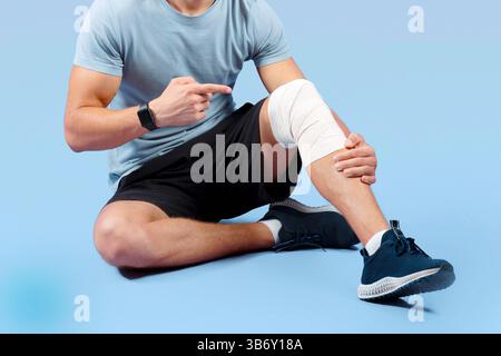 Athletic Millennial Mann mit Sportverletzung, zeigt auf seine verstauchten banded Knie über Flieder Studio Hintergrund Stockfoto