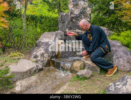 Junger Mann in Anzug des chinesischen (Tibet) buddhistischen Klosters und Wasserquelle im chinesischen Garten (oder japanischen Garten) und er genießt Wasser Stockfoto