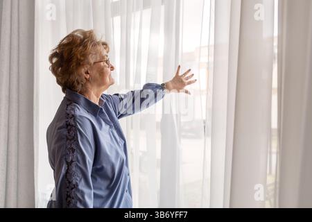 Seniorin in hellblauer Kleidung, die neben dem Fenster steht und sanft durch transparente Vorhänge nach draußen blickt, ein nachdenklicher und friedlicher Moment in einem gemütlichen Zuhause Stockfoto