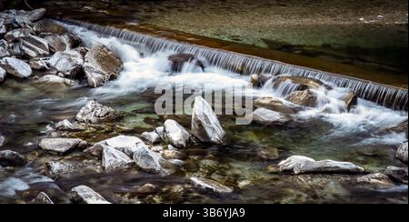 Eine ruhige Flussszene mit fließendem Wasser über Felsen, die eine sanfte Kaskade bilden. Das klare Wasser zeigt Kieselsteine darunter, umgeben von glattem ston Stockfoto