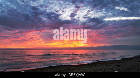 Ein atemberaubender Sonnenuntergang über dem Meer, mit leuchtenden Farben von Orange, Rosa und Lila, die sich auf dem Wasser spiegeln. Der Himmel ist voller dramatischer Wolken, Stockfoto