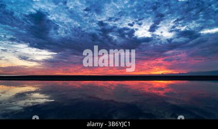 Ein atemberaubender Sonnenuntergang über einem ruhigen Gewässer, der leuchtende Farben am Himmel reflektiert. Die Szene zeigt dramatische Wolken und eine ruhige Landschaft, die kreiert Stockfoto
