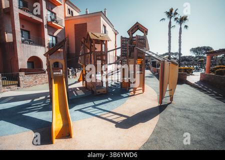 Farbenfroher Spielplatz im Freien mit Holzkletterstrukturen, Seilbrücken und gelben Rutschen in einem sonnigen Innenhof neben einem mediterranen Re Stockfoto