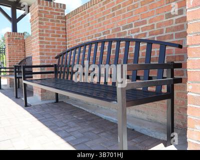 Schwarze Metallbänke mit Lattensitzen und Rückenlehnen, die an Ziegelsteinpflastern in der Nähe von roten Ziegeln in traditionellem Mauerwerk verankert sind, mit einem Blick aus der Nähe von Th Stockfoto
