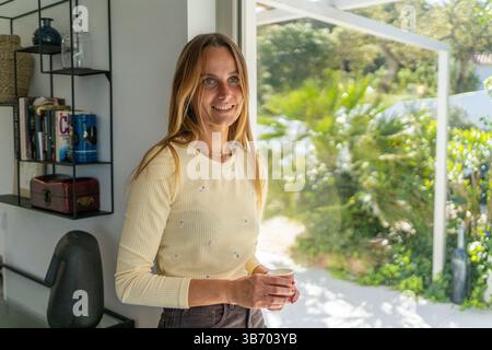 Frau zu Hause, die glücklich lächelt und den Tag beginnt, mit einer Kaffeetasse auf der Hand. Guten Morgen, ich fühle mich ruhig Stockfoto