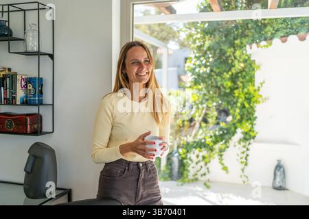 Frau zu Hause am Fenster, beginnt den neuen Tag, lacht mit Tee- oder Kaffeetasse auf der Hand. Wohlbefinden genießen, guten Morgen, glücklich, entspannend Stockfoto