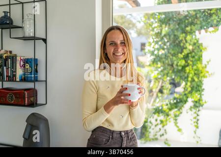 Frau am Fenster zu Hause lacht, beginnt den neuen Tag, genießt Wohlbefinden mit Tee- oder Kaffeetasse auf der Hand. Guten Morgen, glücklich, entspannend o Stockfoto