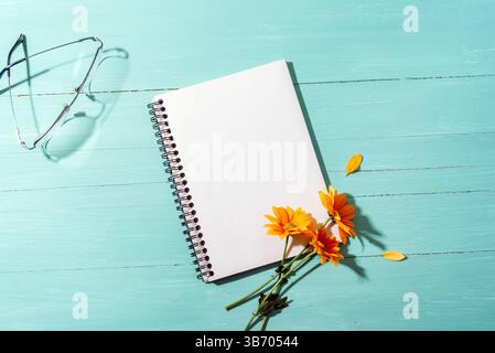 Leeres Spiralnotizbuch mit orangefarbenen Blumen und Brille auf türkisfarbenem hölzernem Hintergrund im Sonnenlicht. Draufsicht, flach, Modell. Stockfoto
