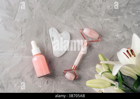 Rosafarbene Serumflasche, Gua Sha Werkzeug und Gesichtsrolle mit weißen Lilienblüten auf grauem strukturiertem Hintergrund. Naturkosmetik, Hautpflegekonzept. Draufsicht, Stockfoto