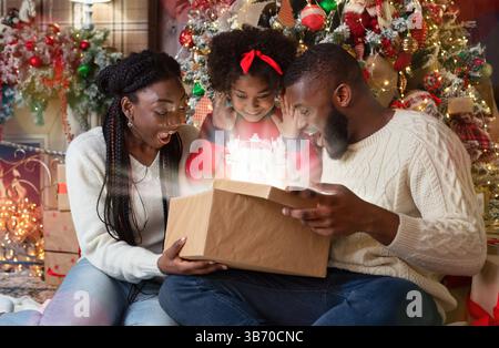 Aufgeregt schwarze Familie von drei Eröffnung scheint Weihnachten Geschenkverpackung Stockfoto