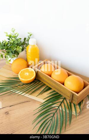 Frische Orangen und Saftflasche auf Holztisch mit grüner Pflanze und Palmblatt vor gelbem Hintergrund Juice Flasche und frische Orangen in Holzkiste Stockfoto