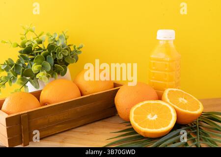Frische Orangen und Saftflasche auf Holztisch mit grüner Pflanze und Palmblatt vor gelbem Hintergrund frische Orangen und Saftflasche auf Holztisch Stockfoto