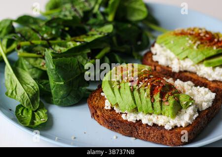 Roggentoast mit Hüttenkäse, Avocado, Sesam, Leinsamen, geräuchertem Paprika und Pfeffer. Serviert mit frischem Spinat, Zitronensaft, Olivenöl und Balsamico Stockfoto