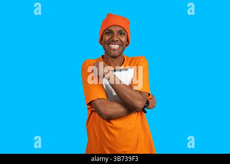 Porträt des glücklichen Liefermannes, der Tablette hält, orangefarbene Uniform und Mütze trägt, lächelnd auf blauem Hintergrund Stockfoto