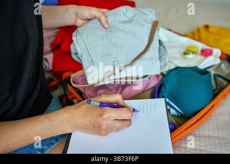 Die Person schreibt in einem Notizbuch mit einem orangen Koffer voller Kleidung. Reisekonzept mit handschriftlicher Checkliste Stockfoto