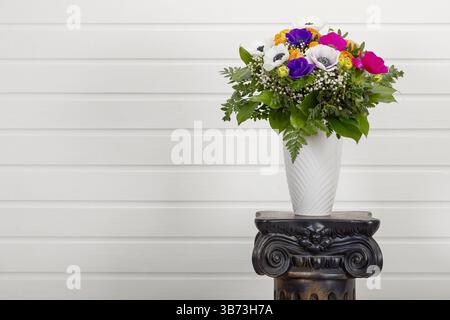 Farbenfrohes Bouquet mit persischen Buttercups und Mohnanemonen in Vase auf antikem Säulensockel mit Kopierraum an weißer Wand Stockfoto
