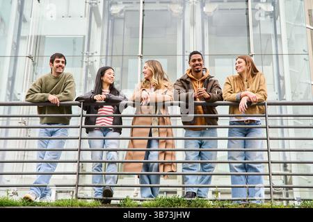 Fünf multiethnische Jugendliche genießen ihre Freizeit, lehnen sich auf ein Geländer vor einem modernen Glasgebäude, plaudern und lächeln. Stockfoto