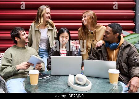 Eine Gruppe verschiedener Studenten arbeitet gemeinsam an Laptops und Tablets in einem Café im Freien, genießt Kaffee und arbeitet an einem Projekt zusammen Stockfoto