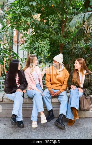 Vier verschiedene Universitätsstudenten, die gemeinsam auf einer Bank in einem Campus-Garten ein Gespräch genießen Stockfoto