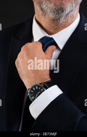 Nahaufnahme eines reiferen Geschäftsmannes, der seine Krawatte anpasst, während er eine klassische Armbanduhr trägt, was Selbstvertrauen und Professionalität symbolisiert. Stockfoto