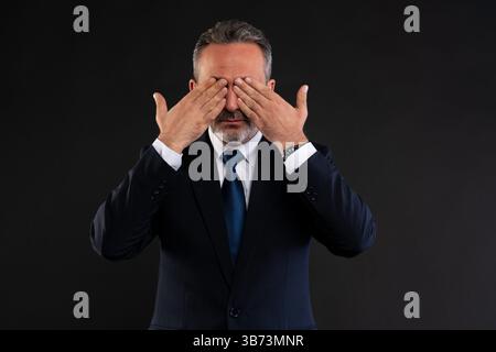 Ein reifer Geschäftsmann im formellen Anzug bedeckt seine Augen mit beiden Händen und deutet auf Verleugnung, Ignoranz oder das Vermeiden der Wahrheit hin. Stockfoto