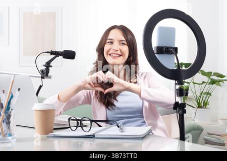 Lächelnde junge weibliche Content Creatorin und Influencerin zu Hause, die mit ihren Händen eine Herzform bildet, um ihren Followern während eines Live-Streamings Liebe zu zeigen Stockfoto