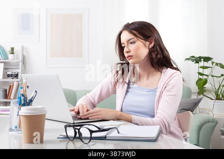 Gelangweilte junge Frau zu Hause mit ihrem Laptop, während sie an einem gemütlichen Schreibtisch sitzt. Darstellung der Remote-Arbeit, des Studiums, des Online-Lernens oder der Nutzung sozialer Medien in einem Stockfoto