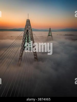 Budapest, Ungarn - Blick aus der Vogelperspektive auf die wunderschöne Megyeri-Hängebrücke über die Donau, die an einem Herbstnachmittag in starkem Nebel bedeckt ist Stockfoto