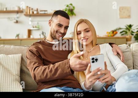 Ein Paar, das dicht auf einer Couch sitzt, lächelt fröhlich zusammen, während es ein Telefon teilt. Stockfoto