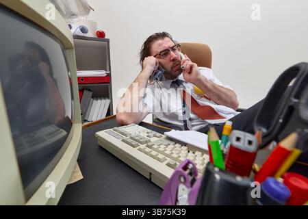 Gestresster Mann, Büroangestellter spricht am Telefon, während er ein Handtuch in Gesicht hält, an einem unordentlichen Schreibtisch sitzt und chaotisches Multitasking und einen Arbeitsplatz zeigt Stockfoto