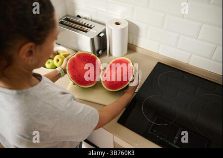 Eine Frau hält zwei Hälften einer frisch geschnittenen Wassermelone in einer hellen, modernen Küche. Die Szene zeigt das häusliche Leben, die Zubereitung frischer Speisen, A Stockfoto