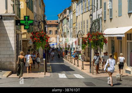 Eine sonnige Straße voller Fußgänger, die an bunten Gebäuden, Geschäften und hängenden Blumenkörben in der Altstadt von Antibes, Frankreich, vorbeilaufen. Stockfoto