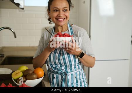 Eine fröhliche Frau genießt eine Schüssel frisches Obst in einer hellen Küche, was einen gesunden Lebensstil fördert. Die farbenfrohen Früchte und die saubere Umgebung sind ein Highlight Stockfoto