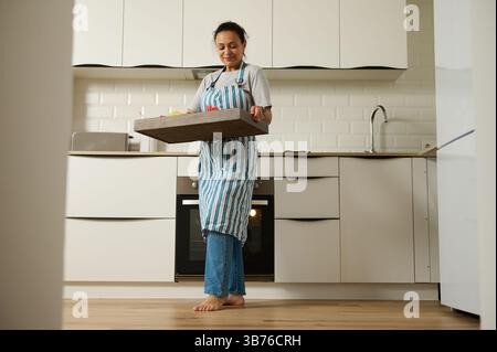 Eine Frau wird in einer modernen Küche beim Zubereiten einer Mahlzeit gesehen. Sie hält ein Tablett und scheint sich auf ihre Aufgabe konzentriert, in einer hellen, sauberen und organisierten Kuppel Stockfoto