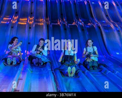 Vier Frauen mit traditionellen bayerischen Dirndls lachen und fangen Selfies auf, während sie auf einer hell beleuchteten Rutsche auf einer lustigen Messe in München am 2. Mai 2025 reiten Stockfoto