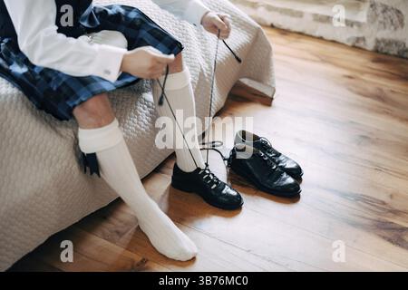 Vorbereitung auf eine schottische Hochzeit. Mann in Kilt, Sporran und hohen Socken sitzt auf dem Bett und bindet lange Schnürsenkel an seine Schuhe. Stockfoto