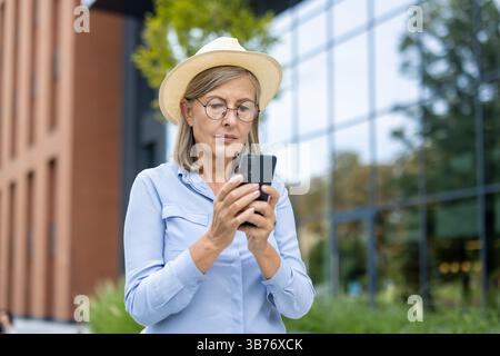 Eine ältere Frau überprüft ihr Handy, während sie an einem sonnigen Tag vor einem modernen Gebäude steht. Sie trägt eine Brille und einen Hut. Stockfoto