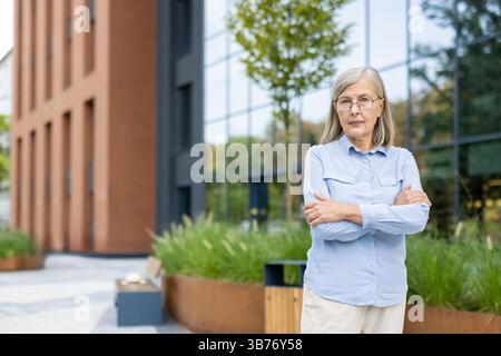 Eine reife Geschäftsfrau steht mit überkreuzten Armen vor einem Bürogebäude und sieht entschlossen aus. Sie trägt eine Brille und ein hellblaues Hemd. Stockfoto