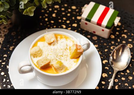 Köstliche Kürbissuppe serviert mit geriebenem Käse und knusprigen Croutons in einer weißen Tasse, perfekt für gemütliche Herbstmahlzeiten. Stockfoto