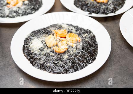 Nahaufnahme eines eleganten Tintenfisch-Risottos, serviert mit Garnelen, Jakobsmuscheln und Parmesan auf einem weißen Teller im Restaurant. Stockfoto