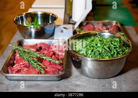 Schüsseln aus Edelstahl gefüllt mit rohen Rindersteaks, frischem Rucola und Kräutern auf einer Küchenzubereitungsstation, die zum Kochen bereit ist. Stockfoto