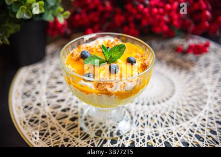 Erfrischendes Mango- und Heidelbeerparfait, garniert mit Schlagsahne, zerbröckelten Keksen und Minzblättern in einem eleganten Glas. Stockfoto