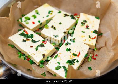 Nahaufnahme von Tofu-Scheiben in einer mit Pergament gefütterten Pfanne, garniert mit gehackten grünen Zwiebeln, Chili und frischen Kräutern. Stockfoto
