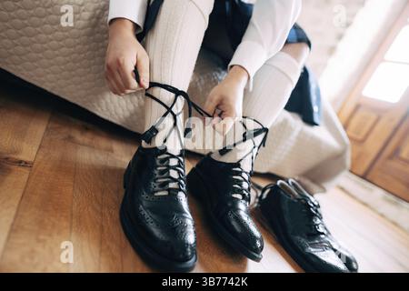 Vorbereitung auf eine schottische Hochzeit. Ein Mann in einem Kilt und hohen Socken sitzt auf dem Bett und bindet lange Schnürsenkel an seinen Schuhen. In der Nähe gibt es ein Paar schwarze Schuhe. Stockfoto