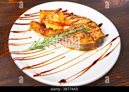 Ein perfekt gegrilltes Lachsfilet wird elegant auf einem weißen Gericht serviert, mit frischem Rosmarin garniert und mit einer reichhaltigen Soße getränkt, was eine Appeal schafft Stockfoto