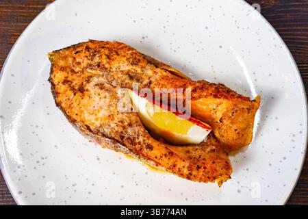 Ein gewürztes Lachsfilet wird perfekt gegrillt und auf einem weißen Teller mit einer Zitronenscheibe für eine erfrischende Note serviert. Ideal für eine gesunde Mahlzeit Opti Stockfoto