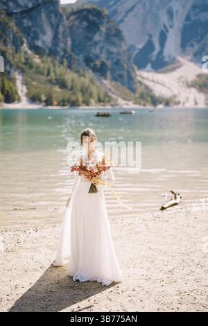 Schöne Braut in einem weißen Kleid mit Ärmeln und Spitze, mit einem gelben Herbststrauß aus getrockneten Blumen und Pfingstrosen, am Pragser Lago in Italien. Stockfoto